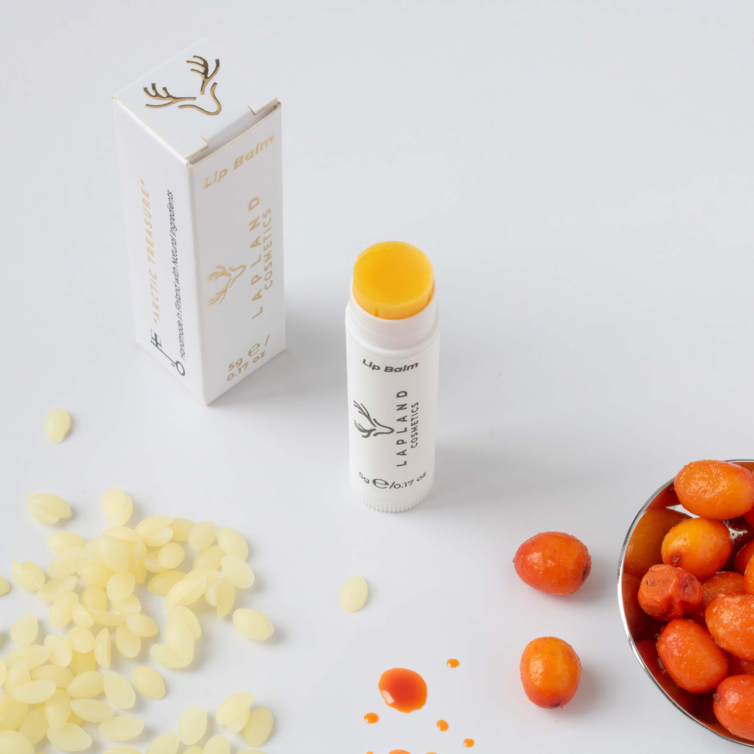 Lapland Cosmetics lip balm with reindeer tallow and Arctic berry oils in a white tube and box, surrounded by beeswax pellets, sea buckthorn berries and oil drops.